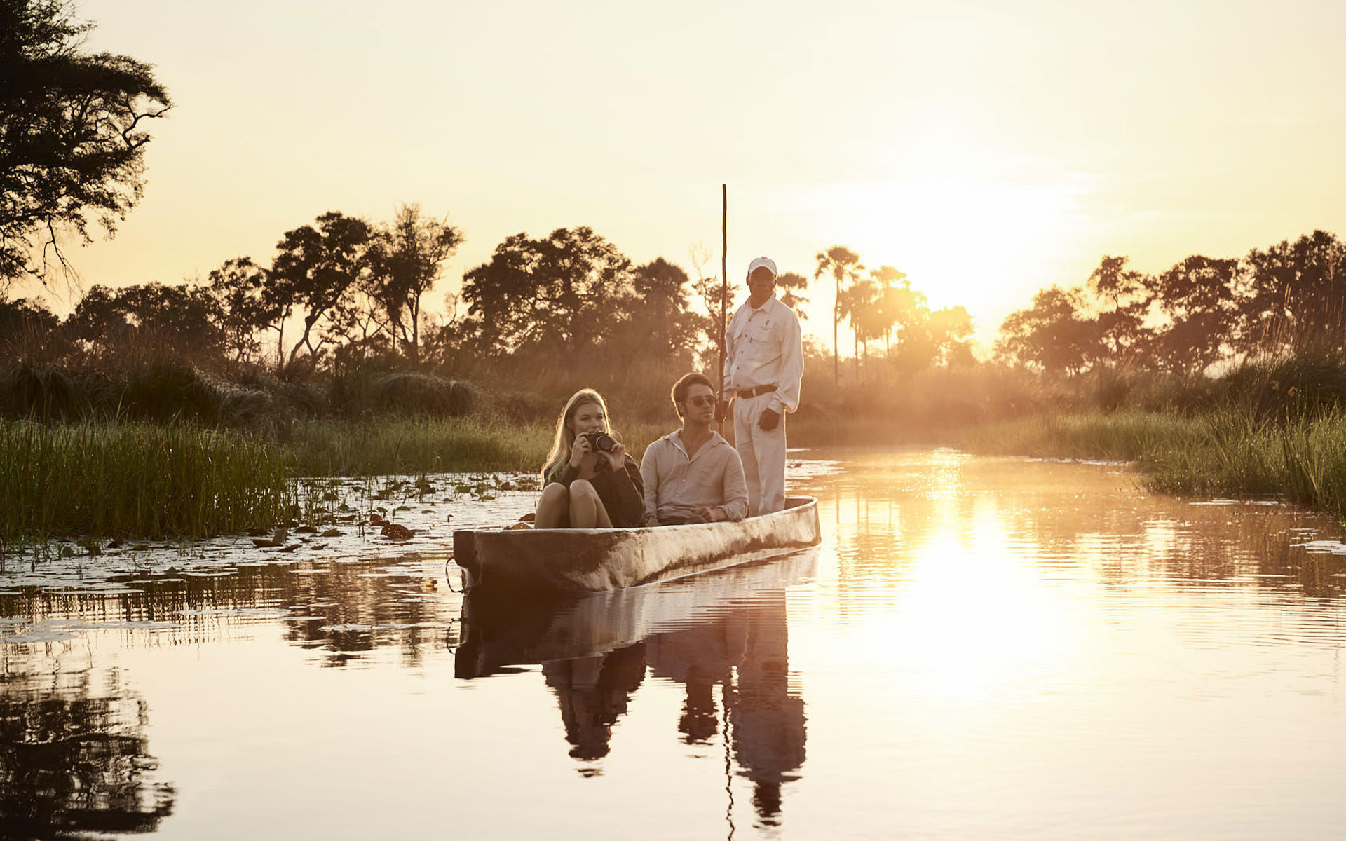 A couple sitting on a mokoro canoe with a poler in the background with Grand Africa Safaris